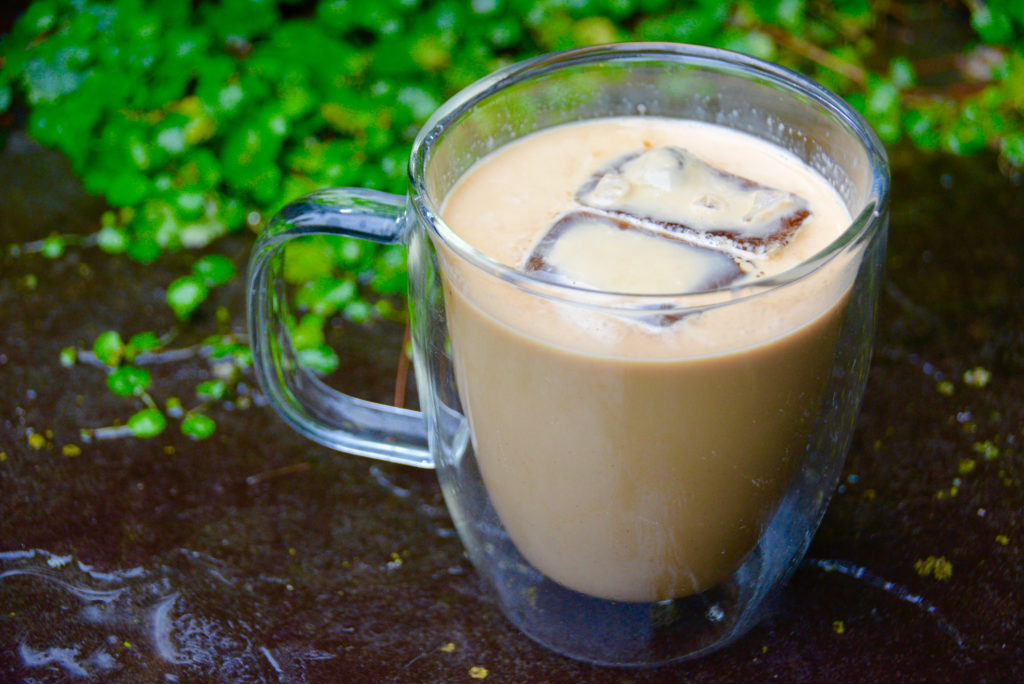 clear mug of iced coffee on pavement with greenery