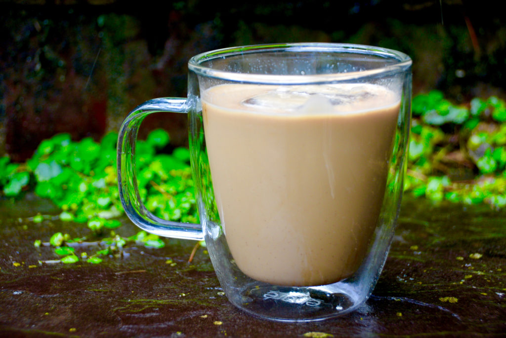 clear mug filled with iced coffee on pavement with greenery in background