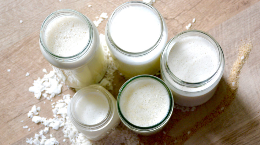 non-dairy milks lined up on a table with coconut shreds, rice grains, and flaxseed