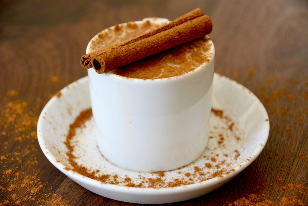 vanilla pumpkin spice latte in a small white coffee mug on a white saucer with cinnamon dusted on top and two cinnamon sticks as garnish