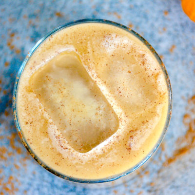 from top - view of pumpkin spice latte in a high ball glass with a blue background a small pumpkin and two ice cubes in the latte