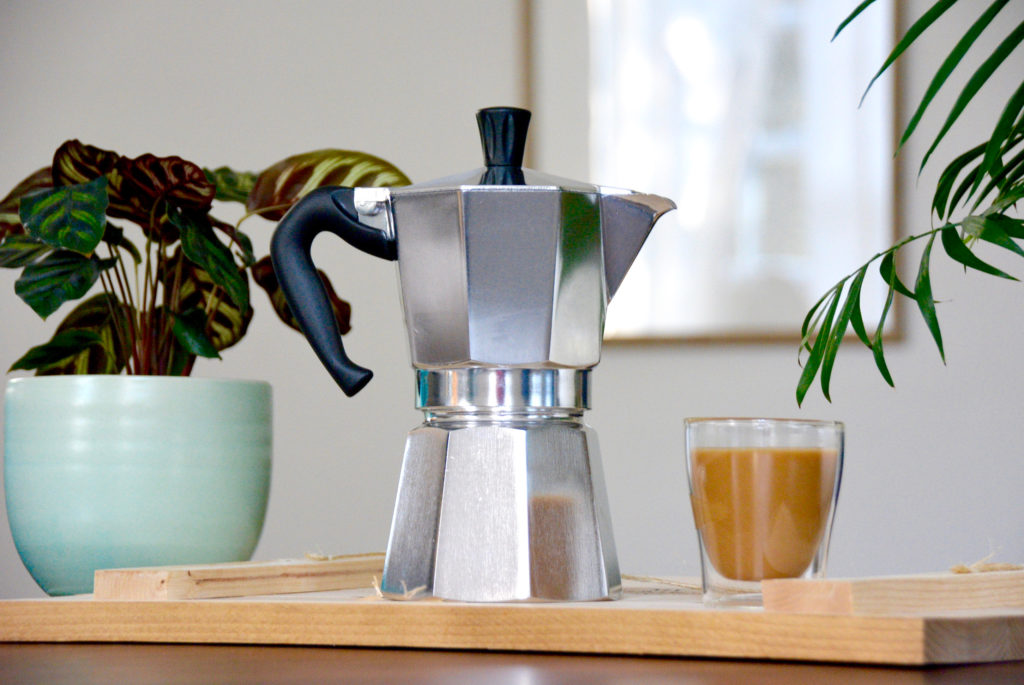 bialetti moka pot next to plants and a cup of coffee on wood table