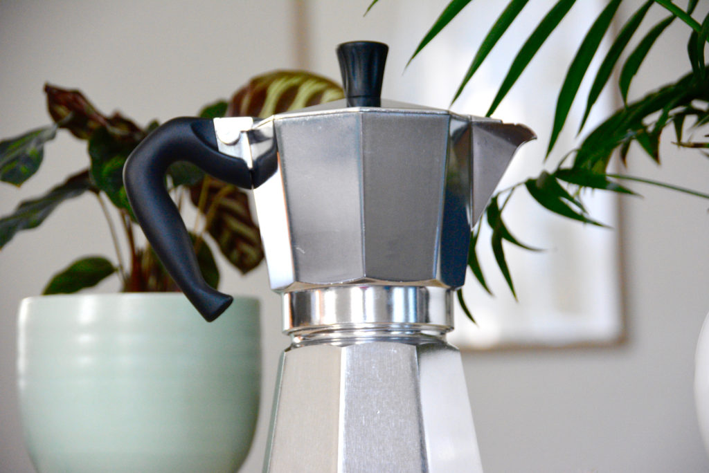 bialetti moka pot next to plants and a cup of coffee on wood table
