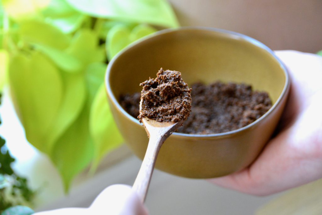 spoon of coffee grounds held over a bowl in front of houseplants 